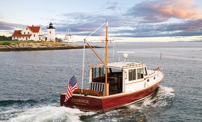 A red hulled lobster yacht cruising past a lighthouse along the coast of Maine