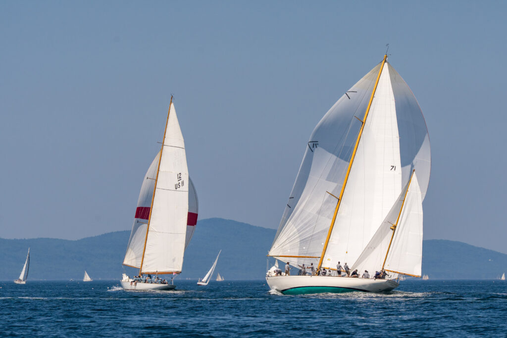 Two sailboats in a race with full sails on blue water with hills on shore in the distance