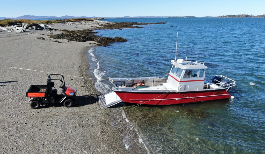 A red aluminum landing craft beached, a four-wheel vehical unloaded on the beach.