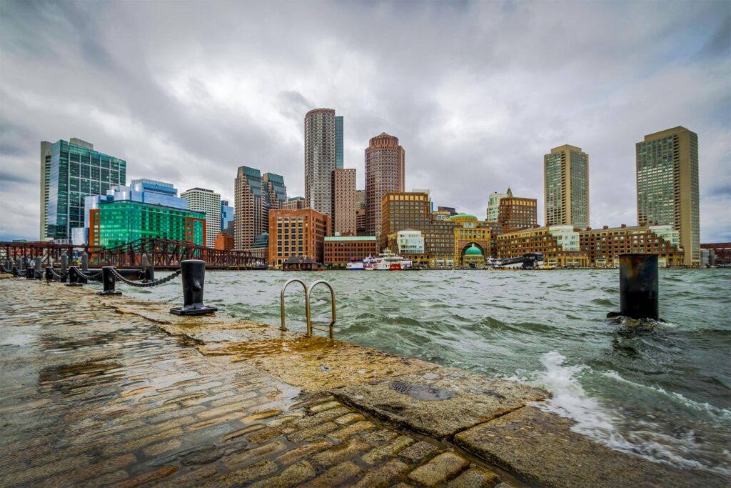 waves crash over the breakwater at high tide, boston skyline in distance