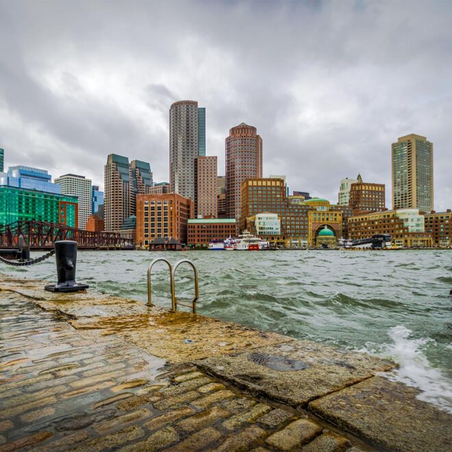 waves crash over the breakwater at high tide, boston skyline in distance