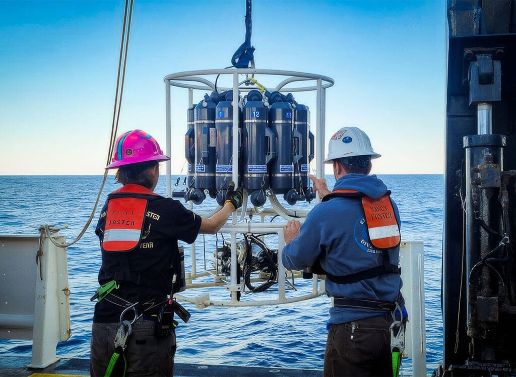 The image depicts two individuals on the deck of a research vessel at sea, engaged in deploying a piece of oceanographic equipment that measures temperature and depth