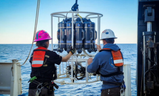 The image depicts two individuals on the deck of a research vessel at sea, engaged in deploying a piece of oceanographic equipment that measures temperature and depth