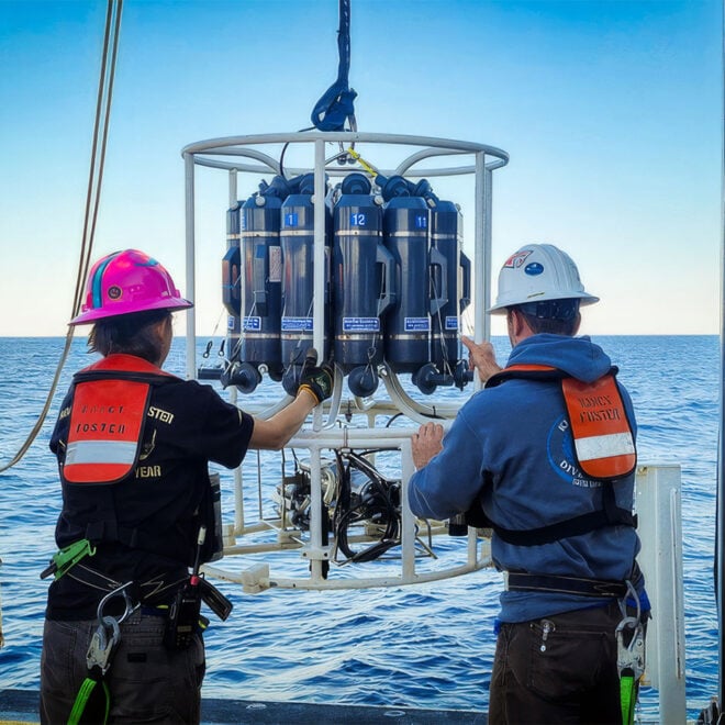 The image depicts two individuals on the deck of a research vessel at sea, engaged in deploying a piece of oceanographic equipment that measures temperature and depth