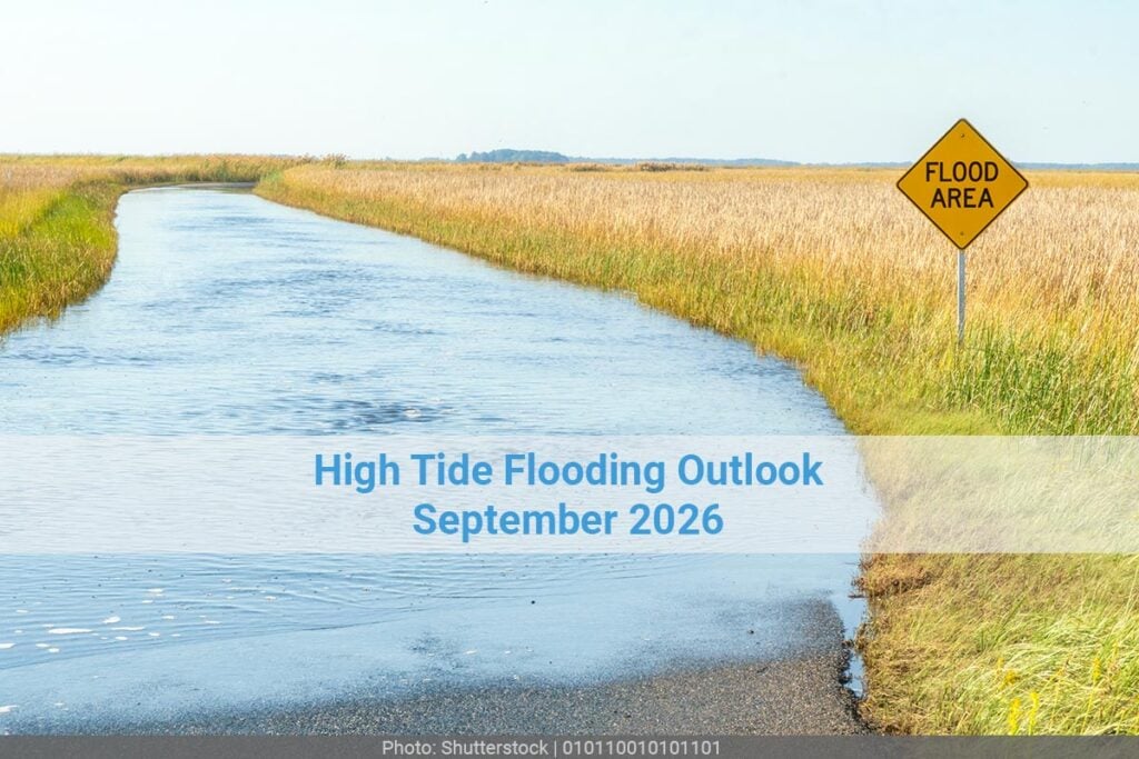 A road sign that reads flood area on a road that is completely flooded and lined by a field on both sides